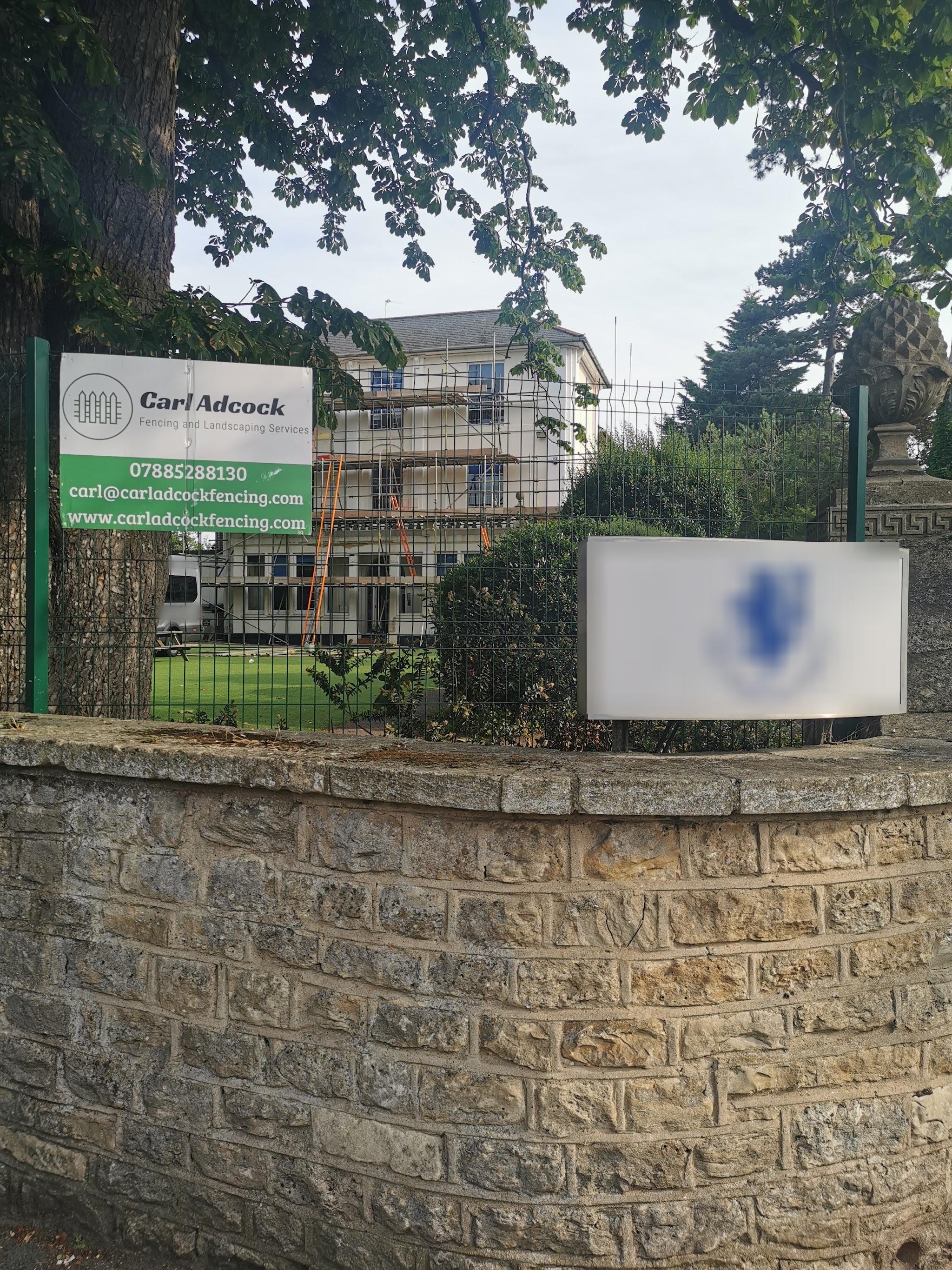 Green mesh security fencing on a stone wall, with the Carl Adcock Fencing site sign